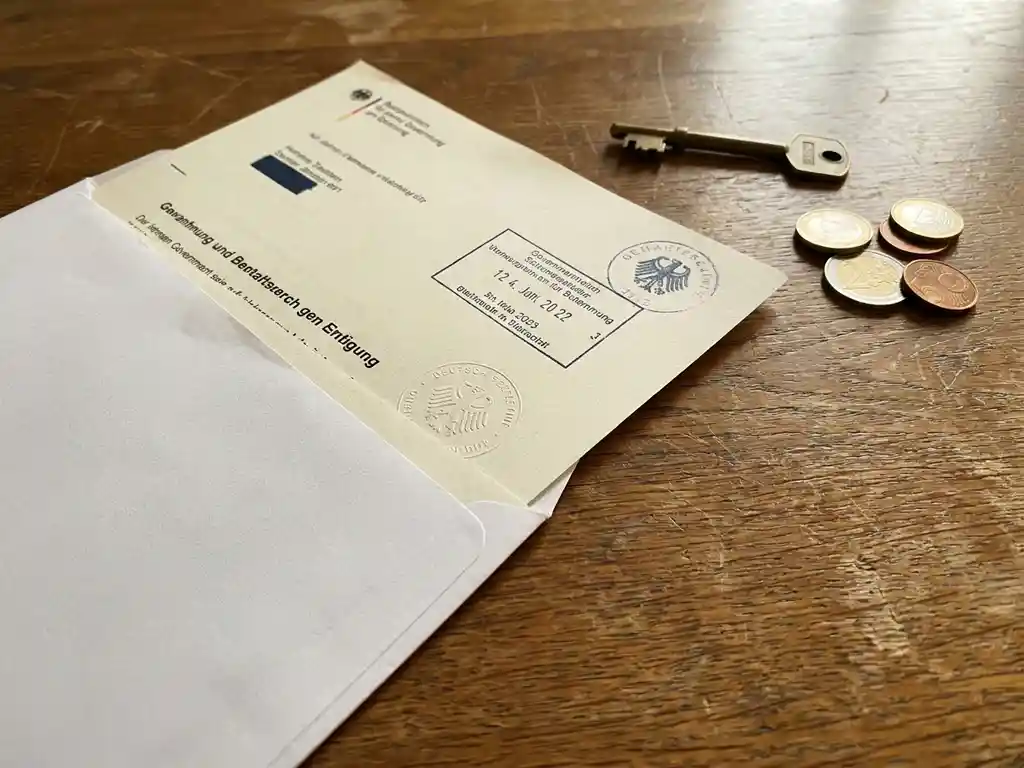 Official German document in white envelope on wooden desk with house key and euro coins in warm afternoon light.